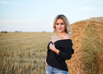 Young blond woman, wearing black jacket and jeans shorts, standing by wheat bale on rural field on summer evening. Three-quarter female portrait during sunset at natural landscape.