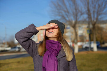 Happy casual excited woman celebrating success stands on the street warmly dressed in a gray hat and a purple. covered her eyes with her hand with happiness or hides her eyes from the bright sun