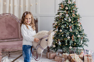 cheerful cute girl playing with a horse on a stick in the living room. Christmas holiday morning. White sweater. Classic light beige tones. Toddler with New Years Gift Toy. Caucasian. Lifestyle child