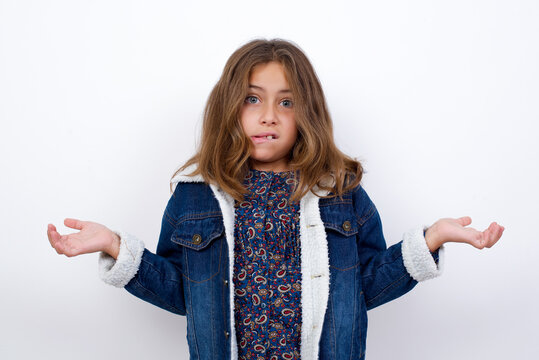 Clueless Little caucasian girl with beautiful blue eyes wearing denim jacket standing over isolated white wall shrugs shoulders with hesitation, faces doubtful situation, spreads palms, Hard decision