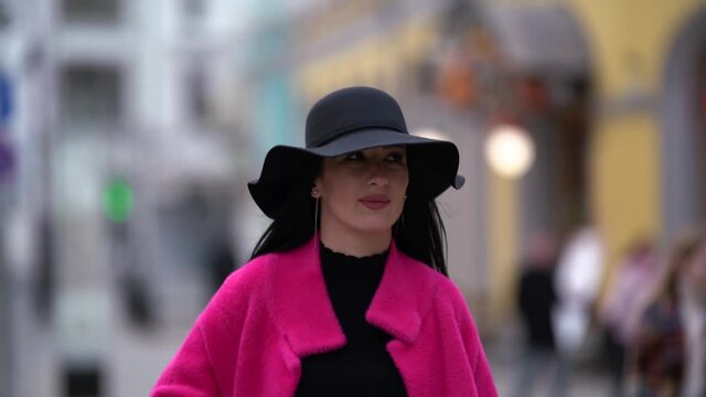 Portrait of a stylish black-haired woman in a pink coat and black hat walking and posing on a blurry background of a busy city street.