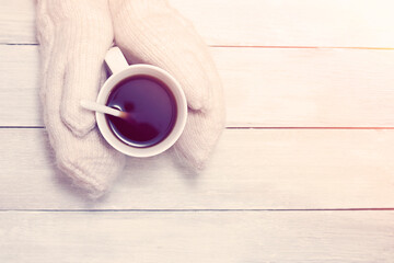 Hands in white knitted mittens holding a cup of hot tea against a white table. New year mood. Christmas scene. Blank for design. Festive layout.