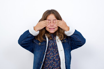 Little caucasian girl with beautiful blue eyes wearing denim jacket standing over isolated white background covering eyes with hands smiling cheerful and funny. Blind concept.