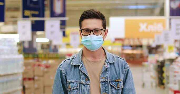 Close up portrait of happy young Caucasian male in mask and glasses standing at supermarket indoors. One handsome cheerful guy customer in food store in quarantine. Virus concept
