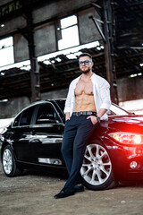 Rich man in unbuttoned white shirt leaned to the luxury car. Black auto at abandoned warehouse.