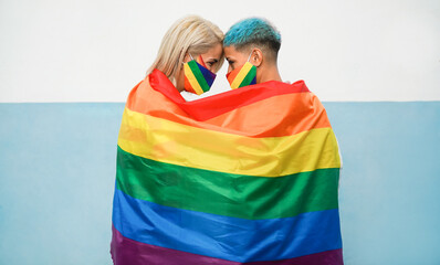 Young couple of women hugging under rainbow flag wearing colorful masks at gay pride event - Lgbt, people and love concept - Focus on faces