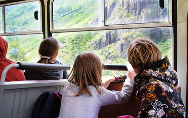 A group of adults and children tourists traveling in a bus looks out the window at the green mountains