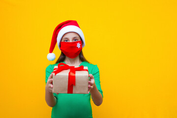 A girl with dark hair in a santa hat in a medical red mask in her hands holds a gift for the holiday Christmas and new year. Isolated yellow background.