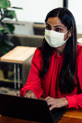Professional Indian American woman working on laptop at business office with mask during pandemic 