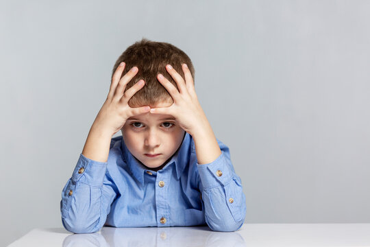Sad schoolboy boy sits at the table and holds his head. Learning difficulties. Gray background. Space for text.