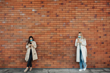 Fototapeta na wymiar Young stylish girls wearing face safety masks are using smartphones, while keeping social distance during coronavirus time. They standing near brick wall.