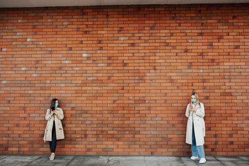 Two women in social distancing standing near brick wall in the city and using phones. Technology and concept of preventing the spread of coronavirus. 