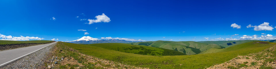 Fototapeta na wymiar Great nature mountain range. Panorama perspective of caucasian mountain or volcano Elbrus with green fields, blue sky background. Elbrus landscape view - the highest peak of Russia and Europe