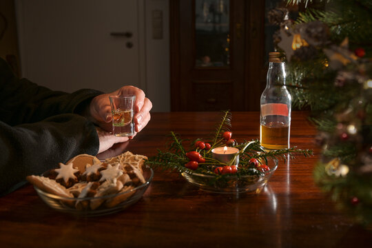 Hands Of An Elderly Man Holding A Shot Glass Alone At A Table With Christmas Decoration, Cookies, Candle And A Half-empty Bottle Of Liquor, Lonely Holidays During The Covid-19 Pandemic, Copy Space