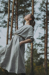 Side view moody portrait of young caucasian woman in a long grey organic linen dress leaning back with face up, eyes closed, enjoying the forest bath in a pine tree forest