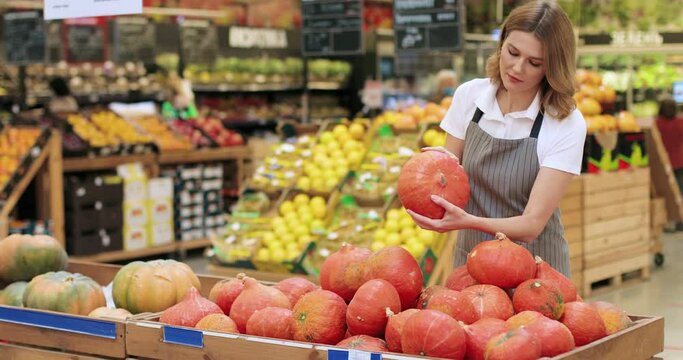 Beautiful female food store worker in apron standing in grocery store and sorting vegetables. Young Caucasian woman employee working indoor and spreads pumpkins at market Woman arranging food products