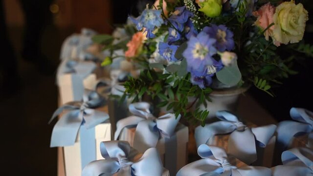 Beautiful festive favors on banquet table with flowers bouquet in the centre, small white paper boxes with blue ribbon prepared for baptism party. Sweet confetti inside the bonbonniere boxes, grateful