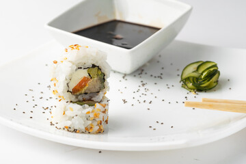 Delicious uramaki sushi with rice, salmon and Philadelphia cheese. Garnished with Cucumber, Poppy Seeds, and Sesame Seeds and a Bowl of Soy Sauce on a White Plate.