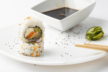 Delicious uramaki sushi with rice, salmon and Philadelphia cheese. Garnished with Cucumber, Poppy Seeds, and Sesame Seeds and a Bowl of Soy Sauce on a White Plate.
