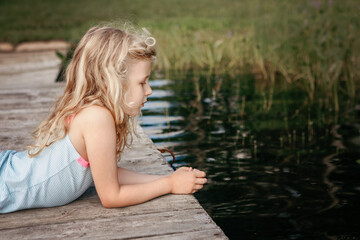 Fototapeta premium Cute pretty Caucasian blonde girl lying on wooden dock pier by lake river and looking in water. Pensive sad thinking child kid explore nature world around. Summer outdoors fun activity.