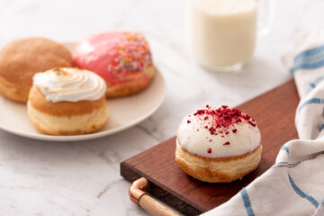 Obraz na płótnie Canvas Close up of a donut with white icing and red decorations on a board with a group of mixed donuts arranged on a plate behind and a glass of milk. Ideal for recipe page or magazine editorial