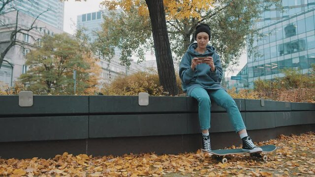 Hipster young woman with skateboard and phone sitting in city park in autumn. Girl playing game on the smartphone . High quality 4k footage