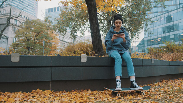 Hipster young woman with skateboard and phone sitting in city park in autumn. Girl playing game on the smartphone . High quality photo