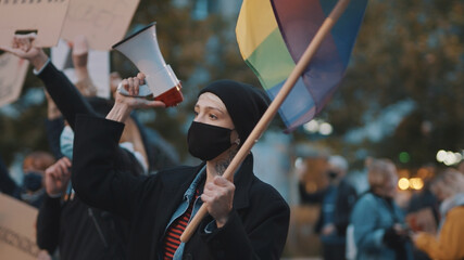 Woman with face mask speaking into the megaphone while holding rainbow flag. Demonstration against...