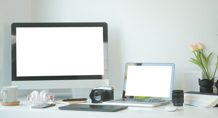 Graphic designer's or photographer workspace with laptop, computer, digital camera, digital tablet and camera accessory on white desk.