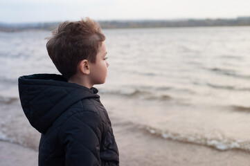 a blond boy in a dark jacket stands in the wind on the Bank of the river and looks into the distance