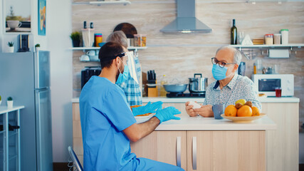 Caregiver holding pills bottle for senior patient during coronavirus pandemic home visit wearing face mask. Male nurse social worker at retired elderly couple explaining covid-19 spreading