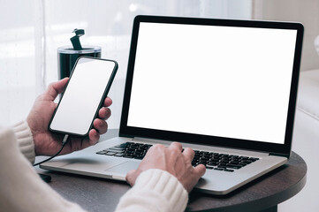 Closeup image of man hands using smartphone and laptop computer while sitting at wooden table at home, blank screen for graphic design.