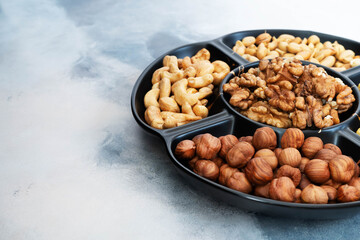 Various shelled nuts (hazelnuts, cashews, walnuts, peanuts) in a round black bowl with five compartments stand on a light gray table. Healthy snack or dessert, vegetarian food. Close-up