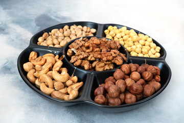 A large serving platter with five compartments in assorted shelled nuts (hazelnuts, cashews, peanuts, walnuts) and chickpeas stands on a light gray concrete background. Healthy food, vitamins