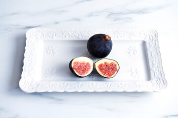 Two ripe fig slices and a fig lie on a white rectangular plate with an embossed pattern. The plate is on a white table. Healthy organic food, seasonal autumn fruits