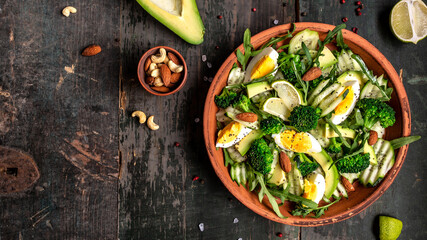 Avocado salad with boiled eggs, broccoli, cucumber, arugula and almond nuts in bowl on dark background. Vegan, detox Buddha bowl, Top view