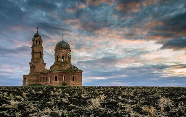 Plowed ground with faded grass, abandoned red brick cathedral against cloudy sky in Edelevo, Russia.