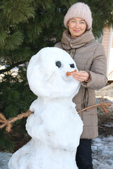 Smiling happy woman wearing warm coat and knitted hat standing near big snowman holding in her hand orange carrot. Winter leisure activity. Vertical, copy space