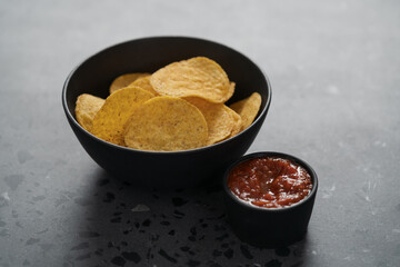 Round nachos in black ceramic bowl on concrete background with red salsa