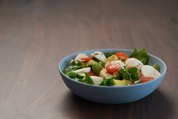 Salad with avocado, cherry tomatoes, romaine and mozzarella in blue bowl on walnut table with copy space