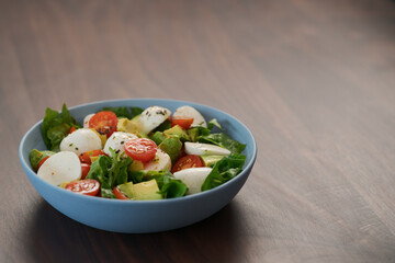 Salad with avocado, cherry tomatoes, romaine and mozzarella in blue bowl on walnut table with copy space