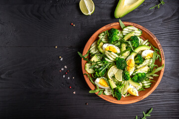 Avocado salad with boiled eggs, broccoli, arugula and almond nuts in bowl on dark background.