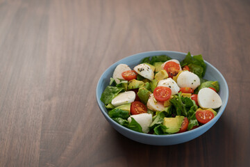 Salad with avocado, cherry tomatoes, romaine and mozzarella in blue bowl on walnut table with copy space