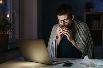 Ill young man having flu, viral infection, suffering from cold symptoms, fever, staying home in self-isolation, take medication, working learning remotely from home at night using laptop at workplace