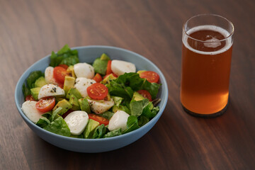 Salad with avocado, cherry tomatoes, romaine and mozzarella in blue bowl and glass of kombucha on walnut table