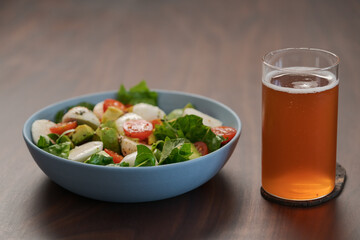 Salad with avocado, cherry tomatoes, romaine and mozzarella in blue bowl and glass of kombucha on walnut table
