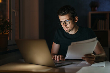 Portrait of tired focused young man in glasses working remotely late at night at home office with...