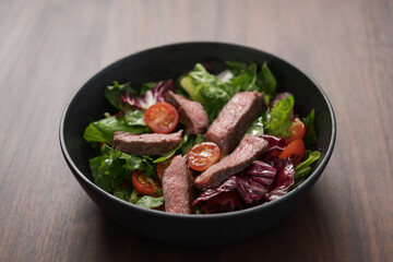 Steak salad with cherry tomatoes and mixed greens in black bowl on walnut wood background