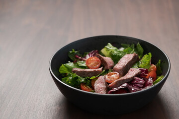 Steak salad with cherry tomatoes and mixed greens in black bowl on walnut wood background