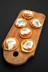Homemade Blini with Smoked Salmon, Creme and Dill on a rustic wooden board on a black surface, low angle view.